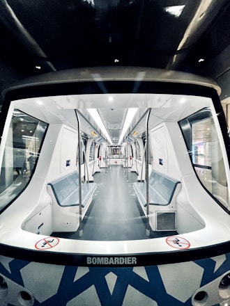 A modern, clean, and empty train interior with sleek, white surfaces and blue accents. The perspective is from the front of the carriage, showing rows of seats lining the walls and metal poles for passengers to hold onto. The ceiling features built-in lighting strips, and there is signage that indicates no standing in certain areas.