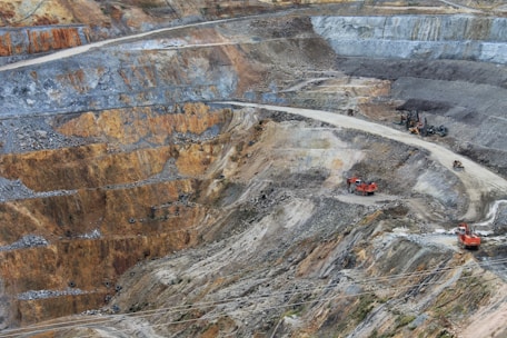 A large open-pit mine with terraced layers of earth and rock. Heavy machinery is visible, including several large red excavators and trucks, scattered across the site. The landscape is marked by various shades of brown, orange, and gray, indicating different layers of soil and rock.