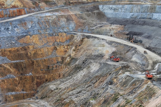 A large open-pit mine with terraced layers of earth and rock. Heavy machinery is visible, including several large red excavators and trucks, scattered across the site. The landscape is marked by various shades of brown, orange, and gray, indicating different layers of soil and rock.
