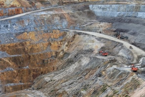 A large open-pit mine with terraced layers of earth and rock. Heavy machinery is visible, including several large red excavators and trucks, scattered across the site. The landscape is marked by various shades of brown, orange, and gray, indicating different layers of soil and rock.