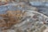 A large open-pit mine with terraced layers of earth and rock. Heavy machinery is visible, including several large red excavators and trucks, scattered across the site. The landscape is marked by various shades of brown, orange, and gray, indicating different layers of soil and rock.