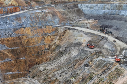 A large open-pit mine with terraced layers of earth and rock. Heavy machinery is visible, including several large red excavators and trucks, scattered across the site. The landscape is marked by various shades of brown, orange, and gray, indicating different layers of soil and rock.