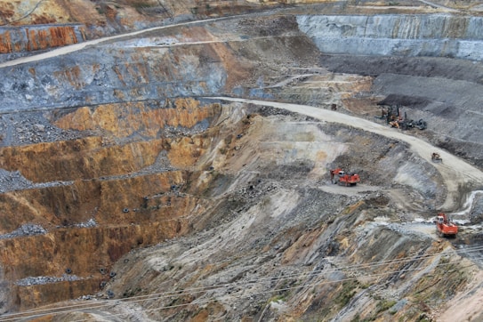 A large open-pit mine with terraced layers of earth and rock. Heavy machinery is visible, including several large red excavators and trucks, scattered across the site. The landscape is marked by various shades of brown, orange, and gray, indicating different layers of soil and rock.