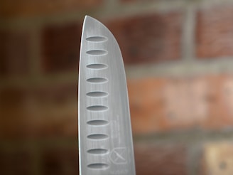 A close-up of the pointed edge of a stainless steel chef's knife, featuring evenly spaced oval-shaped indents along the blade. The background displays a blurred view of a red brick wall.