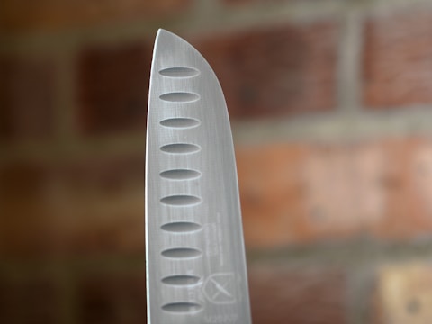 A close-up of the pointed edge of a stainless steel chef's knife, featuring evenly spaced oval-shaped indents along the blade. The background displays a blurred view of a red brick wall.