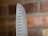 A close-up of the pointed edge of a stainless steel chef's knife, featuring evenly spaced oval-shaped indents along the blade. The background displays a blurred view of a red brick wall.