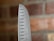 A close-up of the pointed edge of a stainless steel chef's knife, featuring evenly spaced oval-shaped indents along the blade. The background displays a blurred view of a red brick wall.