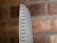 A close-up of the pointed edge of a stainless steel chef's knife, featuring evenly spaced oval-shaped indents along the blade. The background displays a blurred view of a red brick wall.