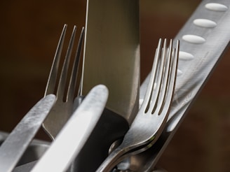 A close-up image featuring several metal kitchen utensils, including forks and a knife, arranged together. The utensils are reflective, displaying a metallic sheen, and the lighting highlights their texture.