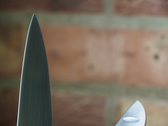 A close-up view of two knife blades with a background of blurred bricks. The focus highlights the sharpness and metallic texture of the blades.