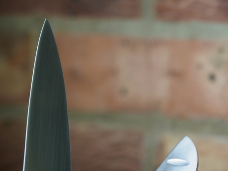 A close-up view of two knife blades with a background of blurred bricks. The focus highlights the sharpness and metallic texture of the blades.