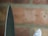 A close-up view of two knife blades with a background of blurred bricks. The focus highlights the sharpness and metallic texture of the blades.
