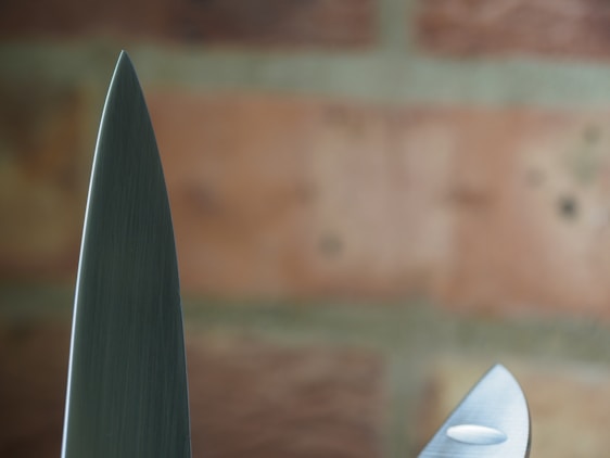 A close-up view of two knife blades with a background of blurred bricks. The focus highlights the sharpness and metallic texture of the blades.