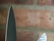 A close-up view of two knife blades with a background of blurred bricks. The focus highlights the sharpness and metallic texture of the blades.