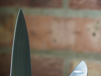 A close-up view of two knife blades with a background of blurred bricks. The focus highlights the sharpness and metallic texture of the blades.