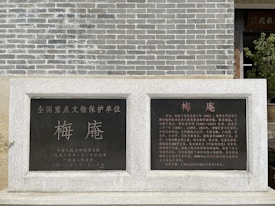A stone monument with Chinese inscriptions in gold and white lettering stands against a gray brick wall. Two black plaques are embedded in the stone, containing text that provides historical information.
