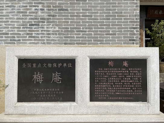 A stone monument with Chinese inscriptions in gold and white lettering stands against a gray brick wall. Two black plaques are embedded in the stone, containing text that provides historical information.