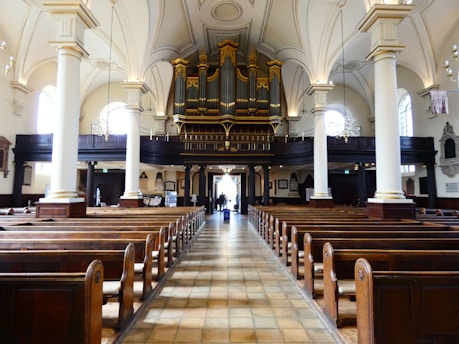 A spacious and ornate church interior featuring rows of wooden pews facing an elaborate pipe organ situated on a balcony. Tall white columns support a vaulted and intricately detailed ceiling. Natural light streams in through large arched windows on the sides, creating a serene and contemplative atmosphere.