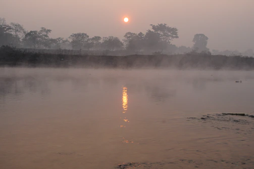 A serene dawn over the Ganges river, with mist rising gently above the water and ancient boats moored along the shore.