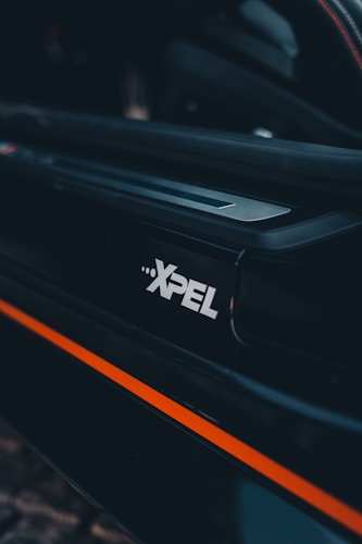 A close-up view of a car door edge featuring the XPEL logo prominently displayed. The design includes a sleek black surface accented with a bold orange stripe. The background suggests an interior car setting with subtle lighting.
