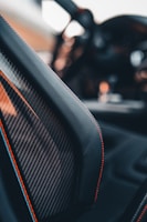 Close-up view of a car interior focusing on a carbon fiber seat with red stitching. The blurred background suggests a luxurious or high-performance vehicle with sleek design elements.
