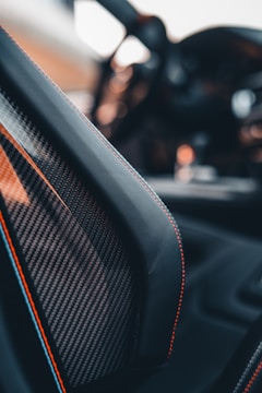 Close-up view of a car interior focusing on a carbon fiber seat with red stitching. The blurred background suggests a luxurious or high-performance vehicle with sleek design elements.