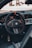 A close-up view of a car's interior focusing on the steering wheel area, which features a sleek design with carbon fiber elements and a black, orange-accented steering wheel. The dashboard displays illuminated gauges and digital screens.