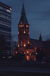 A brick church with a tall steeple and illuminated windows set against a dark evening sky. Nearby, a modern office building displays the logos in bright contrast. Car light trails suggest some traffic on the road in front of the structures.