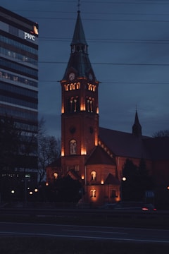 A brick church with a tall steeple and illuminated windows set against a dark evening sky. Nearby, a modern office building displays the logos in bright contrast. Car light trails suggest some traffic on the road in front of the structures.
