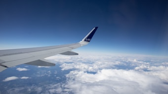 An airplane wing extends into a clear blue sky, with fluffy white clouds visible below. The wing features the letters 'SAS' on the fin.