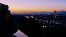 A picturesque view of a cityscape at sunset, featuring a prominent tower in the distance and a softly lit skyline. The sky is a gradient of warm colors, transitioning from orange to deep blue. Silhouettes of people are visible in the foreground, standing on a viewpoint overlooking the city.