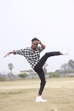 A person is performing a dynamic dance move or martial arts pose in an open field, wearing a black and white checkered shirt, black pants, and white sneakers. The individual is balancing on one leg with the other leg extended high, and wearing sunglasses.
