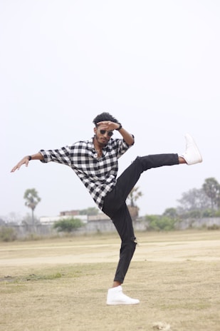 A person is performing a dynamic dance move or martial arts pose in an open field, wearing a black and white checkered shirt, black pants, and white sneakers. The individual is balancing on one leg with the other leg extended high, and wearing sunglasses.