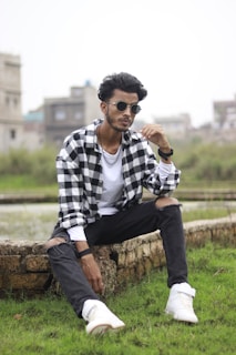 A man wearing sunglasses and casual urban clothing, including a black and white plaid shirt, sits on a stone ledge in a grassy area. He is wearing torn black jeans and white sneakers, with buildings in the blurred background.