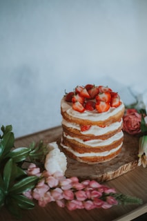 A rustic layer cake topped with fresh strawberries sits on a wooden surface. The cake is partly decorated with cream between layers. Surrounding the cake are various flowers, including pink and white blossoms, adding a touch of elegance to the presentation.