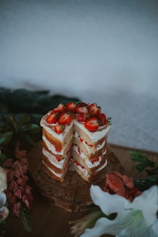 A tall layered cake with white frosting and fresh strawberries on top, set on a wooden slab. The cake appears to be surrounded by various flowers and greenery, creating an elegant presentation.