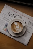 A cappuccino in a white cup sits on top of a folded newspaper. The foam has intricate latte art, including a smooth, swirling pattern. The newspaper is titled 'Le Monde diplomatique' and is arranged on a wooden surface.