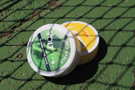 Two round containers are placed on a textured green surface. The container in the foreground has a green label with text, while the one partially visible in the background has a yellow label. A shadow pattern from a net falls across both containers.