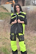 A person is standing outdoors, wearing a neon yellow and black work outfit. There is construction equipment, such as an excavator, in the background, and the area appears to be a construction site with dirt and sparse vegetation.