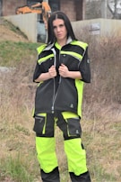 A person is standing outdoors, wearing a neon yellow and black work outfit. There is construction equipment, such as an excavator, in the background, and the area appears to be a construction site with dirt and sparse vegetation.
