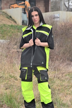 A person is standing outdoors, wearing a neon yellow and black work outfit. There is construction equipment, such as an excavator, in the background, and the area appears to be a construction site with dirt and sparse vegetation.