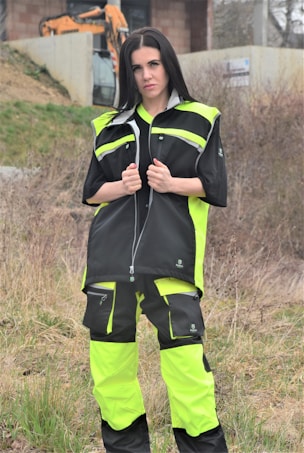 A person is standing outdoors, wearing a neon yellow and black work outfit. There is construction equipment, such as an excavator, in the background, and the area appears to be a construction site with dirt and sparse vegetation.