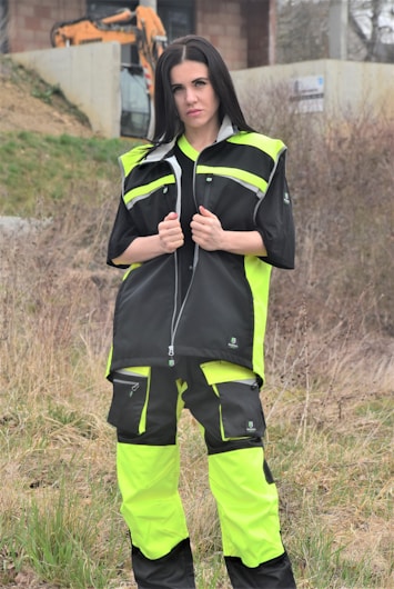 A person is standing outdoors, wearing a neon yellow and black work outfit. There is construction equipment, such as an excavator, in the background, and the area appears to be a construction site with dirt and sparse vegetation.