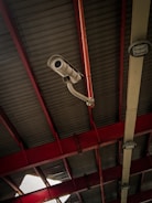 A security camera is mounted on a metal beam inside a building with a high ceiling. The ceiling is composed of corrugated metal panels supported by red structural beams. Two lights are also visible on either side of the camera, attached to the beams. A section of the ceiling shows damage, allowing light to shine through.