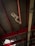 A security camera is mounted on a metal beam inside a building with a high ceiling. The ceiling is composed of corrugated metal panels supported by red structural beams. Two lights are also visible on either side of the camera, attached to the beams. A section of the ceiling shows damage, allowing light to shine through.