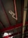 A security camera is mounted on a metal beam inside a building with a high ceiling. The ceiling is composed of corrugated metal panels supported by red structural beams. Two lights are also visible on either side of the camera, attached to the beams. A section of the ceiling shows damage, allowing light to shine through.