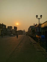 A quiet city street at sunset with a warm orange glow in the sky. The sun is low on the horizon, casting long shadows on the empty road. Street lamps line the sidewalk, and a few vehicles and people are visible in the distance. Buildings on either side are partially illuminated by the setting sun.