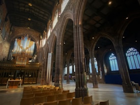 A large, ornate church interior with high vaulted ceilings and tall arches. The scene is dominated by a grand organ at one end, illuminated by light streaming through stained glass windows. Rows of wooden chairs are lined up neatly in the foreground. The architecture is intricate, with detailed carvings and stone columns creating a majestic and solemn atmosphere.