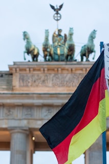A German flag waves prominently in the foreground, with the historic Brandenburg Gate in the background, crowned by the Quadriga statue of a chariot drawn by four horses.