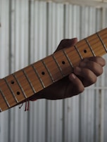 A hand is gripping the neck of a guitar, pressing down on the strings. The guitar has a wooden neck with metal frets and dot markers. The background is a corrugated metal surface, creating a contrast with the wooden texture of the guitar.